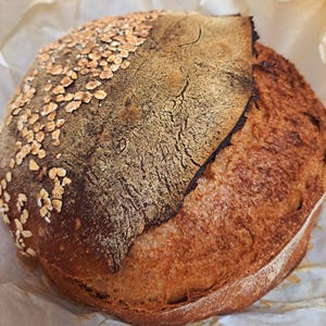 Könnte beinhalten: Ein rundes Brot aus handwerklicher Herstellung mit goldbrauner Kruste. Die Oberseite ist teilweise mit Hafer bedeckt. Die Kruste hat ein strukturiertes Aussehen und das Brot liegt auf Backpapier.