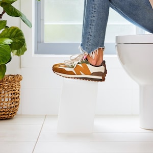May include: A brown, white, and green athletic shoe with a white sole. The shoe is on a white surface. The person is wearing blue jeans with distressed knees and frayed hems. A green plant and a woven basket are in the background.