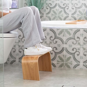 May include: A curved wooden step stool in a bathroom. The stool is made of light-coloured wood and has a simple, modern design. The stool is approximately 30 cm tall. The background features patterned tiles and a bathtub.