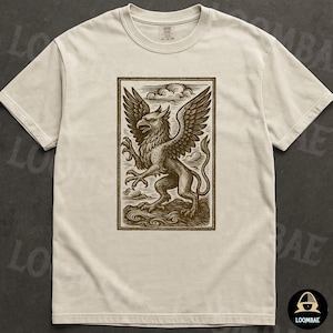 May include: Cream-colored t-shirt featuring a brown illustration of a griffin within a rectangular frame. The griffin, a mythical creature with an eagle's head and wings and a lion's body, is depicted with detailed linework. The shirt has a crew neck and short sleeves.