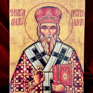 May include: An icon depicting a religious figure with a long white beard, wearing a red and gold crown and robes. The figure holds a red book and is set against a gold background with Cyrillic text. The artwork is framed.