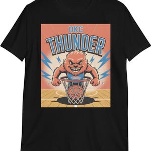 Op de afbeelding: Zwart T-shirt met een cartoon illustratie van een beer die een basketbal dunk. De beer is oranje en blauw, met de tekst "OKC THUNDER" erboven. De achtergrond is oranje met blauwe bliksemschichten.