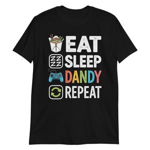 May include: Black t-shirt with the words "EAT SLEEP DANDY REPEAT" in white, with colorful accents. The design includes a takeout container, Zzz's, a game controller, and a recycle symbol. A fun graphic tee.