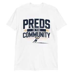 Può includere: T-shirt bianca con la scritta "PREDS IN THE COMMUNITY" in blu navy e oro. Sotto il testo c'è una grafica di un giocatore di hockey. La maglietta è realizzata in un materiale morbido e confortevole.