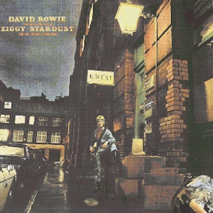 Può includere: Una copertina di album quadrata che mostra una persona in piedi in un vicolo buio e bagnato di notte. Il testo recita DAVID BOWIE e ZIGGY STARDUST. Un cartello sull'edificio in mattoni riporta K. WEST. Un lampione illumina la scena con luce gialla.