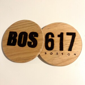 May include: Two round wooden coasters with black lettering. One coaster says "BOS" and the other says "617 BOSTON".