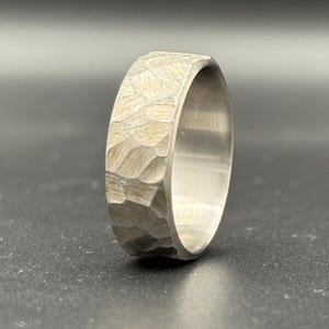 Op de afbeelding: Een zilveren ring met een gehammerde textuur. De ring is breed en heeft een afgerond profiel.
