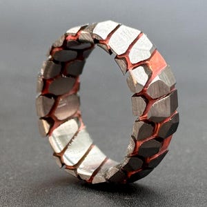 May include: A silver ring with a textured, geometric design. The ring has a red inlay that creates a contrasting pattern.