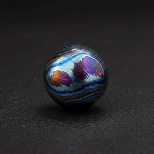 May include: A spherical object with a metallic, iridescent finish. The surface displays a blend of blues, purples, and oranges, creating a marbled effect. The object is set against a dark background, highlighting its vibrant colors and unique texture.
