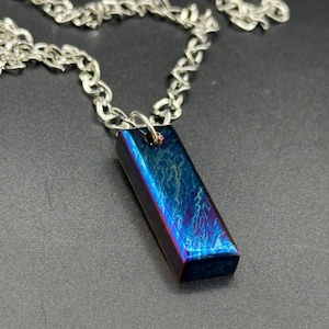 Könnte beinhalten: Halskette mit rechteckigem Anhänger und silberner Kette. Der Anhänger hat ein blau-violettes schillerndes Finish. Die Kette ist silberfarben. Die Halskette wird auf einer dunkelgrauen Oberfläche präsentiert.