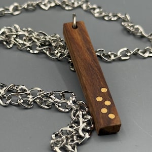 Puede incluir: Un collar con colgante de madera con una cadena plateada. El colgante rectangular es marrón con cuatro puntos dorados. La cadena plateada es una cadena de eslabones.
