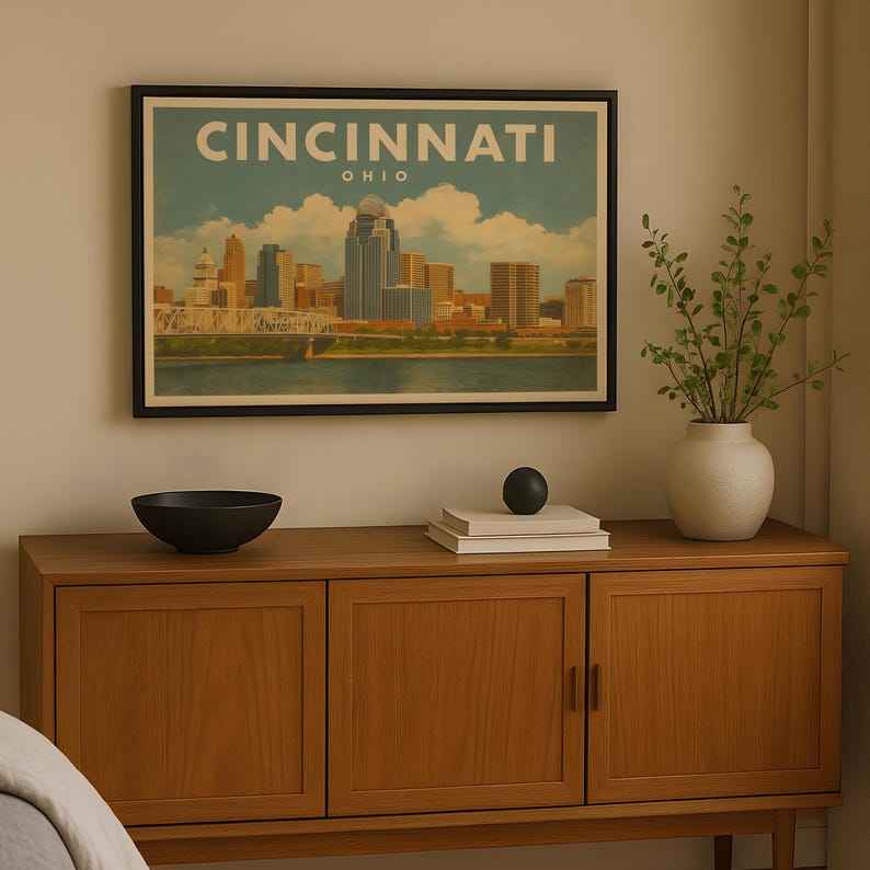 May include: A framed print of the Cincinnati, Ohio skyline, with the city name in vintage lettering. The artwork shows the cityscape with a river and cloudy sky. The print is displayed above a wooden cabinet with a black bowl and decorative items.
