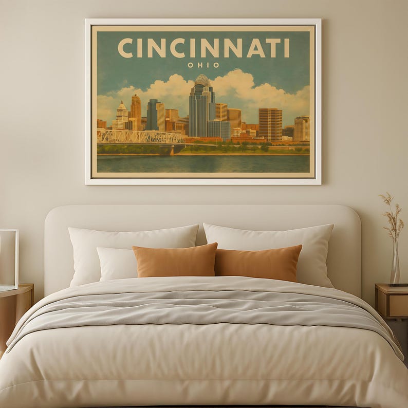 May include: A framed print of the Cincinnati, Ohio skyline in a vintage travel poster style. The artwork is displayed above a bed with neutral-toned bedding. The city name is at the top of the print.