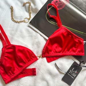 Puede incluir: Conjunto de lencería de satén rojo con tirantes ajustables y detalles de cadena dorada. El conjunto incluye un sujetador y una braguita. También es visible una etiqueta negra con el texto "LA TOM Atelier de Lingerie".