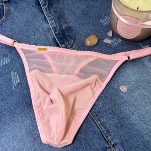 Puede incluir: Lingerie rosa transparente con un delicado ribete de encaje y herrajes dorados. La lencería tiene un corte de talle bajo y una espalda de tanga.