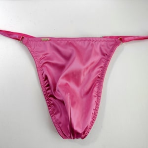 Puede incluir: Ropa interior de satén rosa con tirantes ajustables. La ropa interior tiene una cintura baja y una parte trasera de tanga.