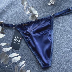 Könnte beinhalten: Ein dunkelblauer Satin-String mit verstellbaren Seitenträgern, die mit silberfarbenen Metallringen und Schiebern versehen sind. Ein schwarzes rechteckiges Etikett ist befestigt und zeigt den weißen Text LIA TOM Atelier de Lingerie mit einem roten Punkt.