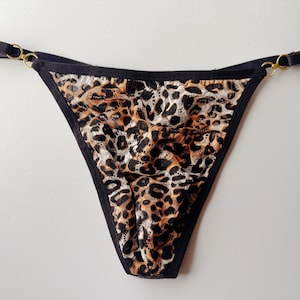 May include: A thong-style undergarment with a leopard print pattern. The fabric is a mix of brown, black, and cream. The edges are trimmed with black fabric, and the straps are black with gold-colored metal rings.