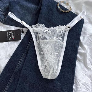 Puede incluir: Tanga de encaje blanco con patrones florales y tirantes ajustables. La lencería se muestra sobre un par de vaqueros azul oscuro. Una etiqueta dice "LIA TOM Atelier de Lingerie". También se ve una pulsera.