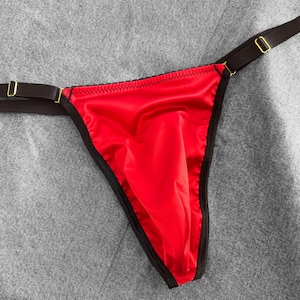 May include: Red satin thong underwear with black trim and straps. The straps are black with gold-colored metal hardware. The underwear is displayed on a gray surface.