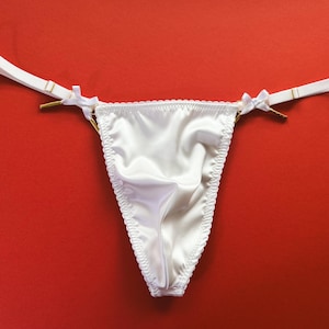 May include: White satin thong underwear with delicate trim and thin straps. The straps are attached with gold-colored hardware and small white bows. The background is a solid red.