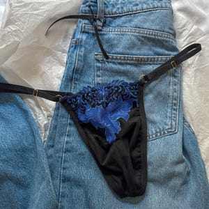 May include: Black thong underwear with blue floral embroidery. The lingerie features thin straps with gold-colored hardware. The garment is displayed on a pair of blue denim jeans.