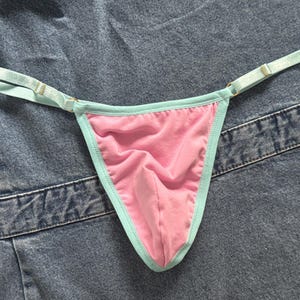 May include: Pink thong underwear with a light blue trim and straps. The straps are adjustable with gold-colored metal hardware. The underwear is made of a soft fabric.