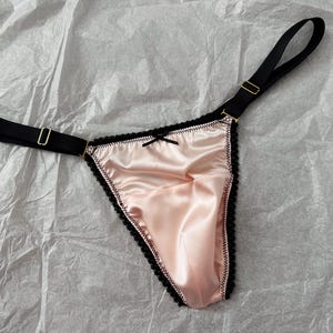 Peut inclure: Un string en satin rose blush avec une bordure noire et un petit nœud. Le string est doté d'une ceinture élastique noire et d'une sangle avec une boucle en métal doré.