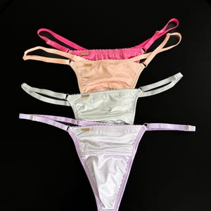 Op de afbeelding: Vier string-slips in tinten roze, perzik, wit en lavendel. Elke string heeft dunne bandjes en een delicaat ontwerp. Het ondergoed is gerangschikt op een zwart oppervlak, waarbij de kleuren en stijlen worden getoond.