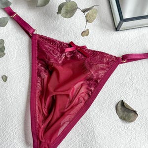 Puede incluir: Tanga burdeos con delicados detalles de encaje y un pequeño lazo. La lencería presenta tirantes ajustables y una tela transparente, creando un look femenino y elegante. Una pieza elegante para una colección de lencería.