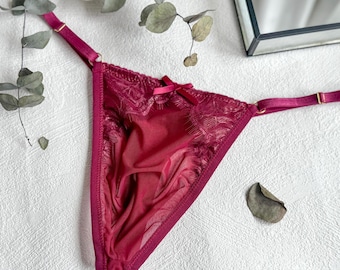 Op de afbeelding: Bordeaux string-slip met delicate kanten details en een kleine strik. Het lingerie heeft verstelbare bandjes en een transparante stof, wat een vrouwelijke en elegante uitstraling creëert. Een stijlvol stuk voor een lingerieset.