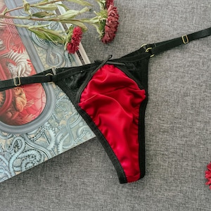 May include: Black and red lingerie set. The thong features a red satin front panel and black lace detailing. Adjustable straps with gold-tone hardware complete the design. A small black bow is at the front. The lingerie is displayed with a book and flowers.