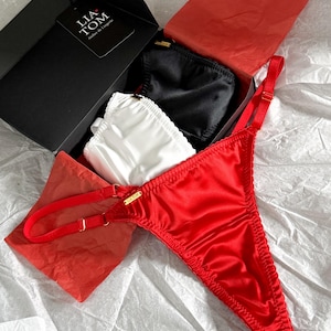 Peut inclure: Un string en satin rouge avec des bretelles réglables est présenté avec une boîte noire contenant de la lingerie blanche et noire. La boîte porte une étiquette « LIA TOM Atelier de Lingerie ». La lingerie est présentée sur une surface blanche texturée.