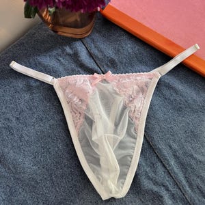 Puede incluir: Tanga blanco y transparente con ribete de encaje rosa y un pequeño lazo. La lencería presenta un diseño delicado y tirantes finos. El fondo incluye una superficie de mezclilla y un jarrón con flores moradas.