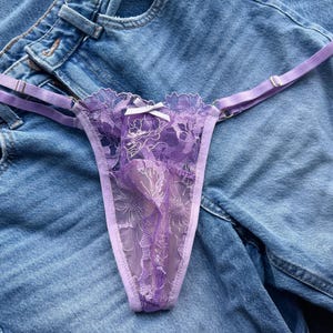 Può includere: Un perizoma delicato color lavanda con dettagli in pizzo floreale e un piccolo fiocco bianco. La lingerie presenta spalline regolabili ed è esposta su uno sfondo in denim.