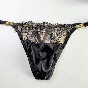 Puede incluir: Lingerie negra de encaje con bordado floral. La lencería tiene un panel trasero de satén y tirantes ajustables.