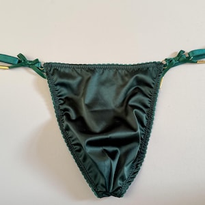 May include: Dark green satin thong underwear with delicate scalloped edges. The straps are a matching green with small bows and gold-colored hardware. A luxurious and elegant lingerie piece.