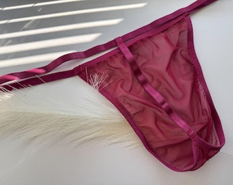 Op de afbeelding: Een diep magenta string gemaakt van transparante mesh-stof met dunne, elastische bandjes met een satijnen afwerking. Het ontwerp bevat een verticaal bandje dat de tailleband met het voorpand verbindt, wat zorgt voor een gestroomlijnde look.