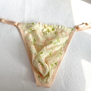 Puede incluir: Tanga crema con estampado floral en rosa y verde. La lencería presenta un diseño de encaje, un pequeño lazo y tirantes ajustables. Los bordes están ribeteados en un tono rosa suave, creando una estética femenina.