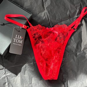 Puede incluir: Un tanga de encaje rojo con tirantes ajustables y herrajes dorados. La lencería se exhibe sobre una superficie oscura junto a una caja negra y una etiqueta que dice "LIA TOM Atelier de Lingerie."
