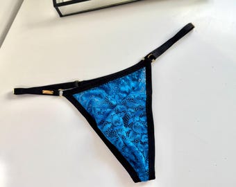 Op de afbeelding: Een blauwe kanten string met zwarte randen en bandjes. De string heeft een bloemen kanten patroon en goudkleurige hardware. Het kledingstuk wordt op een wit oppervlak gepresenteerd.