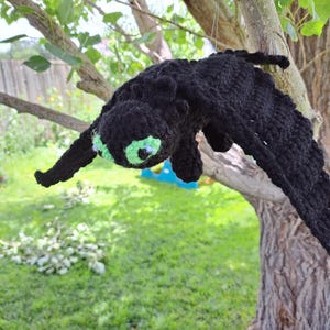 May include: A black crocheted dragon with green eyes and wings is suspended from a tree branch. The handmade dragon has a detailed texture and is set against a backdrop of green grass and foliage.