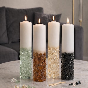 May include: Four lit pillar candles with a decorative base of colored glass fragments. The candles are white with bases in green, brown, gold, and black. Matches and loose glass fragments are on the table.