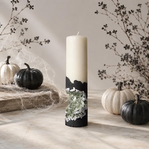 May include: A cylindrical pillar candle with a cream-colored top and a black base. The black section features a textured, jagged cutout revealing translucent, pale green crystalline structures embedded within the wax surface.