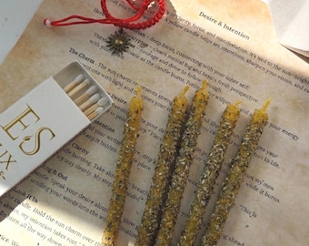 May include: An overhead shot features yellow candles with a textured, speckled design, alongside a matchbox and a red bracelet. The candles are arranged on a paper with text. The scene evokes a spiritual or ritualistic theme.