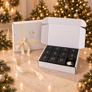 May include: A white gift box with a cream ribbon and the gold text ES LUX CANDLE, next to an open white box containing twelve black square boxes numbered one through twelve in gold, with a small round candle revealed under the number twelve.