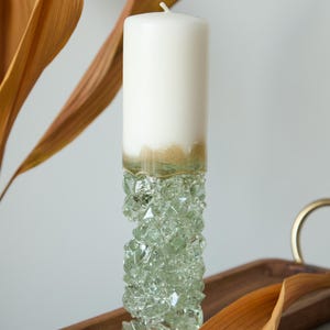 Op de afbeelding: Een witte kaars met een gouden rand staat op een heldere glazen kaarsenhouder gemaakt van gestapelde kristal-achtige stukken. De kaarsenhouder staat op een houten dienblad met gedroogde bladeren.