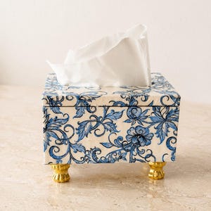 May include: A square tissue box cover featuring a white base with a blue floral and vine pattern. The box has a removable lid and is supported by four small, gold-colored feet with a textured, claw-like design. A white tissue protrudes from the top.