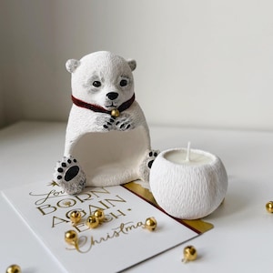 May include: A white polar bear figurine with a red collar and a gold bell, holding a small white candle. A greeting card with gold text and several gold bells are in the foreground. The card reads "Love Born Again Each Christmas."