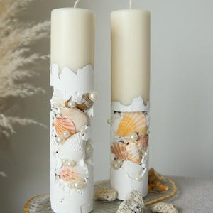 May include: Two cylindrical off-white pillar candles decorated with white textured material, assorted seashells in shades of peach, tan, and white, and small white faux pearls. The candles are displayed on a clear oval tray with gold trim.